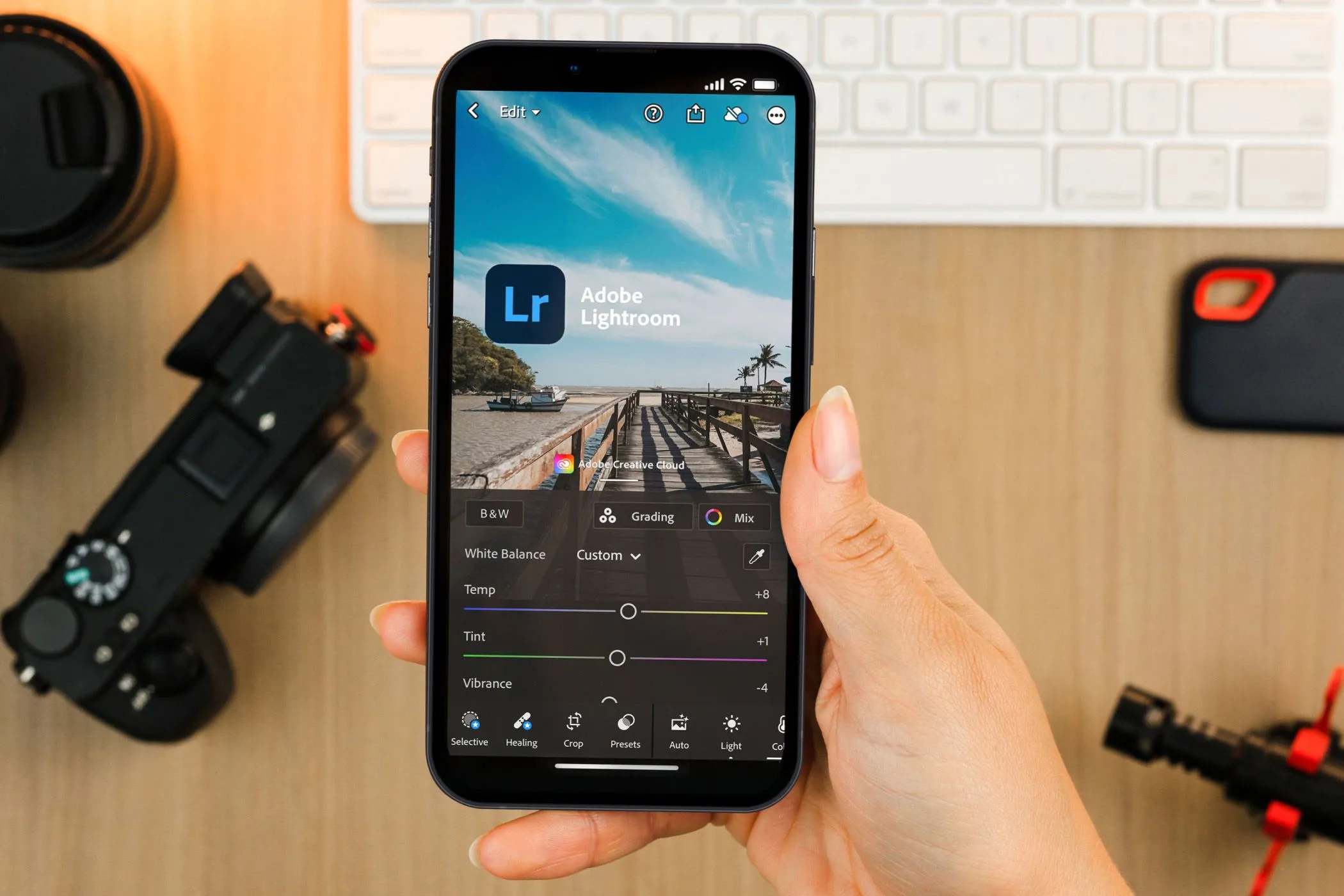 Một người đang cầm điện thoại hiển thị ứng dụng Lightroom Mobile, với thiết bị chụp ảnh chuyên nghiệp ở hậu cảnh.