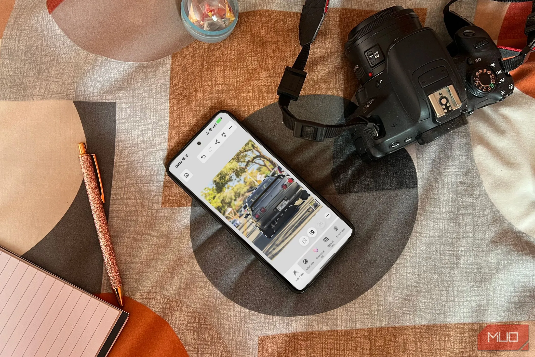 Người dùng đang trải nghiệm ứng dụng Photoshop trên điện thoại Android màu đen đặt trên giường cạnh máy ảnh và sổ tay